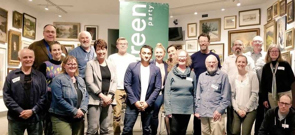 A group of Green Party candidates and volunteers gathered in front of a Green Party banner at an art gallery.